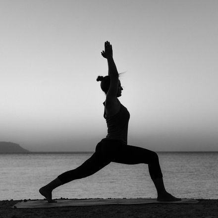 Woman in Warrior pose on a yoga mat at the beach, arms raised, black and white.