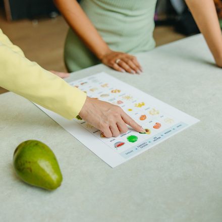 Two people examining a chart with fruit samples, one points at an avocado.