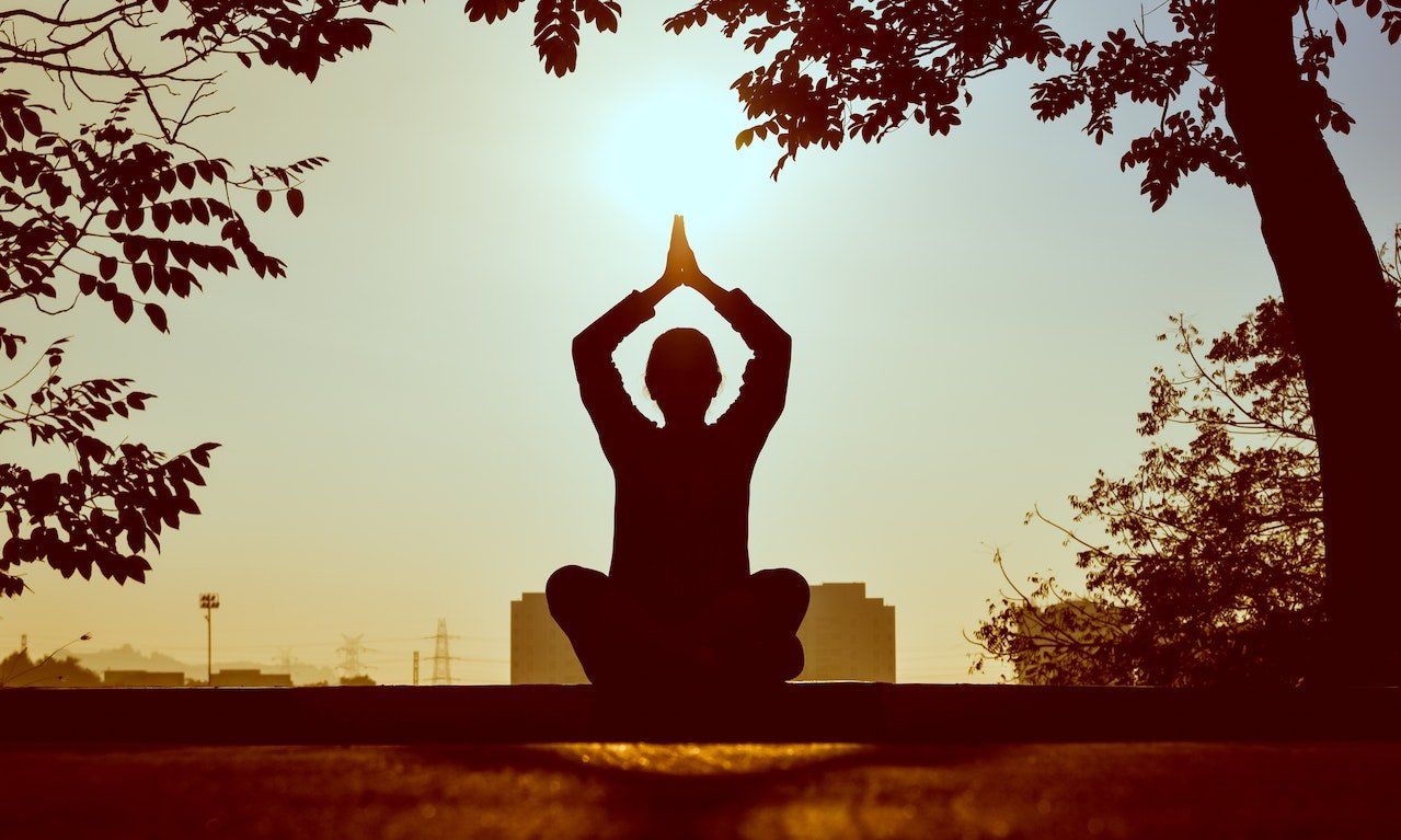 Silhouette of person in lotus position, hands in prayer pose, with sun and tree branches in the background.