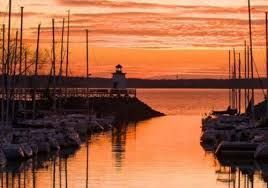 Sunset over a marina with sailboats, calm water, and a small lighthouse on the pier