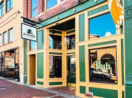 Colorful storefront with green and yellow trim, large windows, and a hanging sign on a brick street.