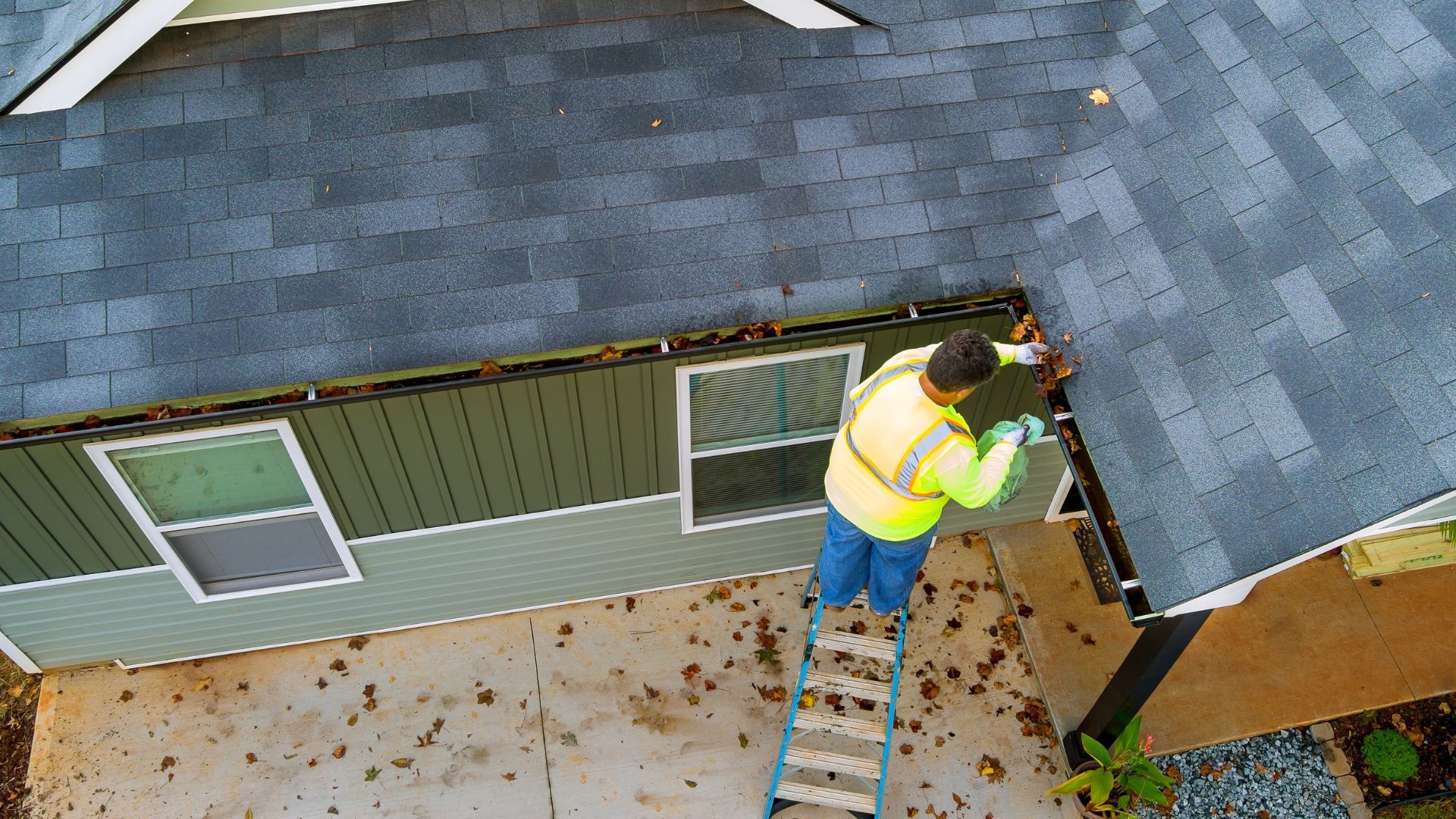 An aerial view of a man on a ladder cleaning a gutter on the side of a house.