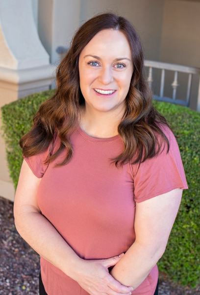 Woman in pink shirt smiles outdoors with arms crossed.