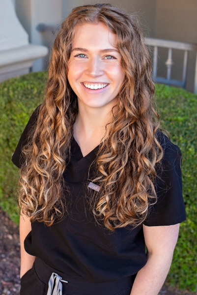Woman with curly brown hair smiling, wearing black scrubs, standing near a trimmed bush and building.