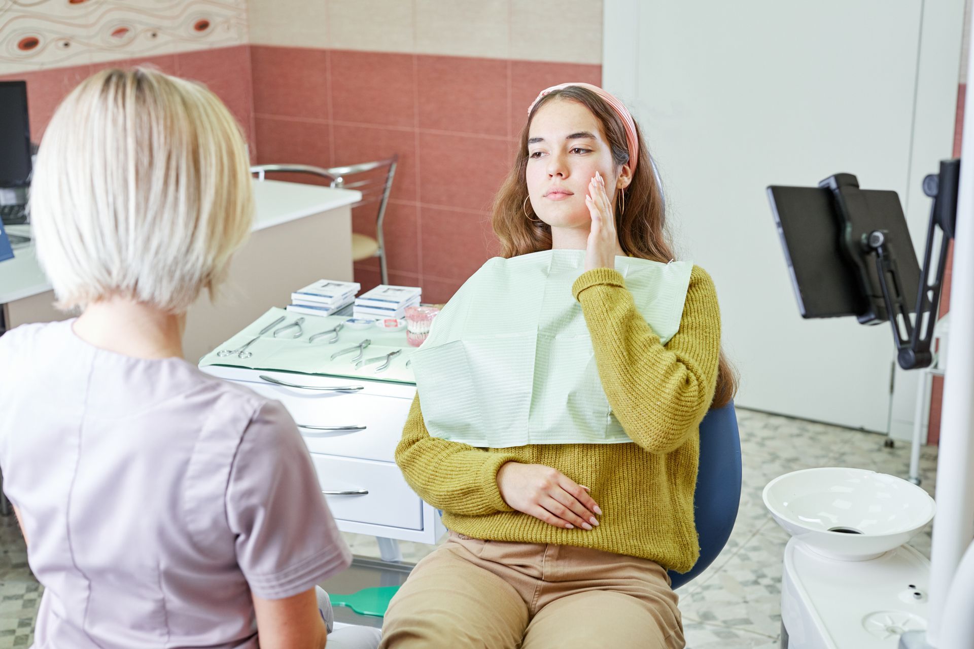 Patient seated in dental chair while clinician speaks in a treatment room, with bib and dental tools visible