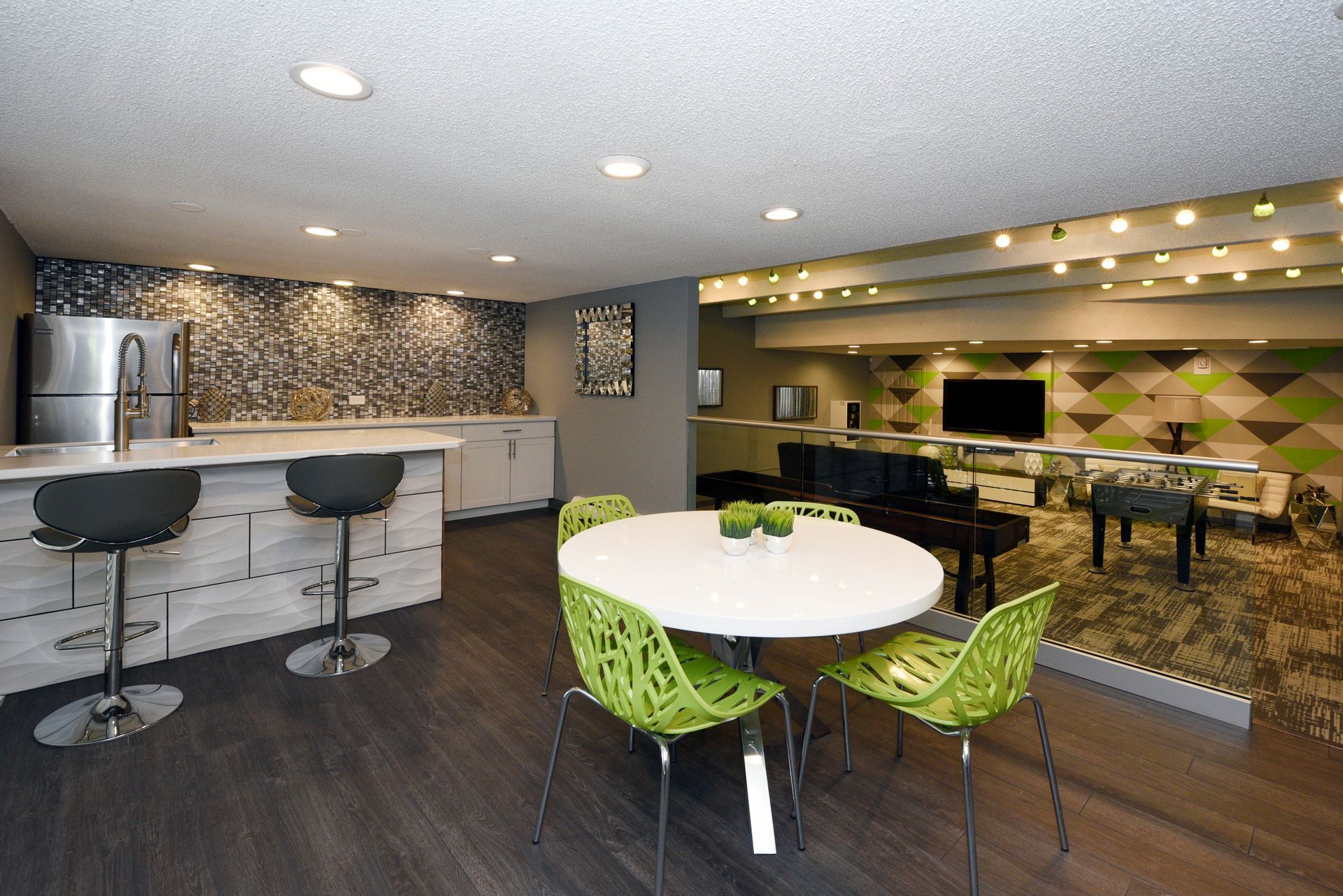 Modern lounge with white tables, green chairs, a bar, and a TV wall in a softly lit interior.