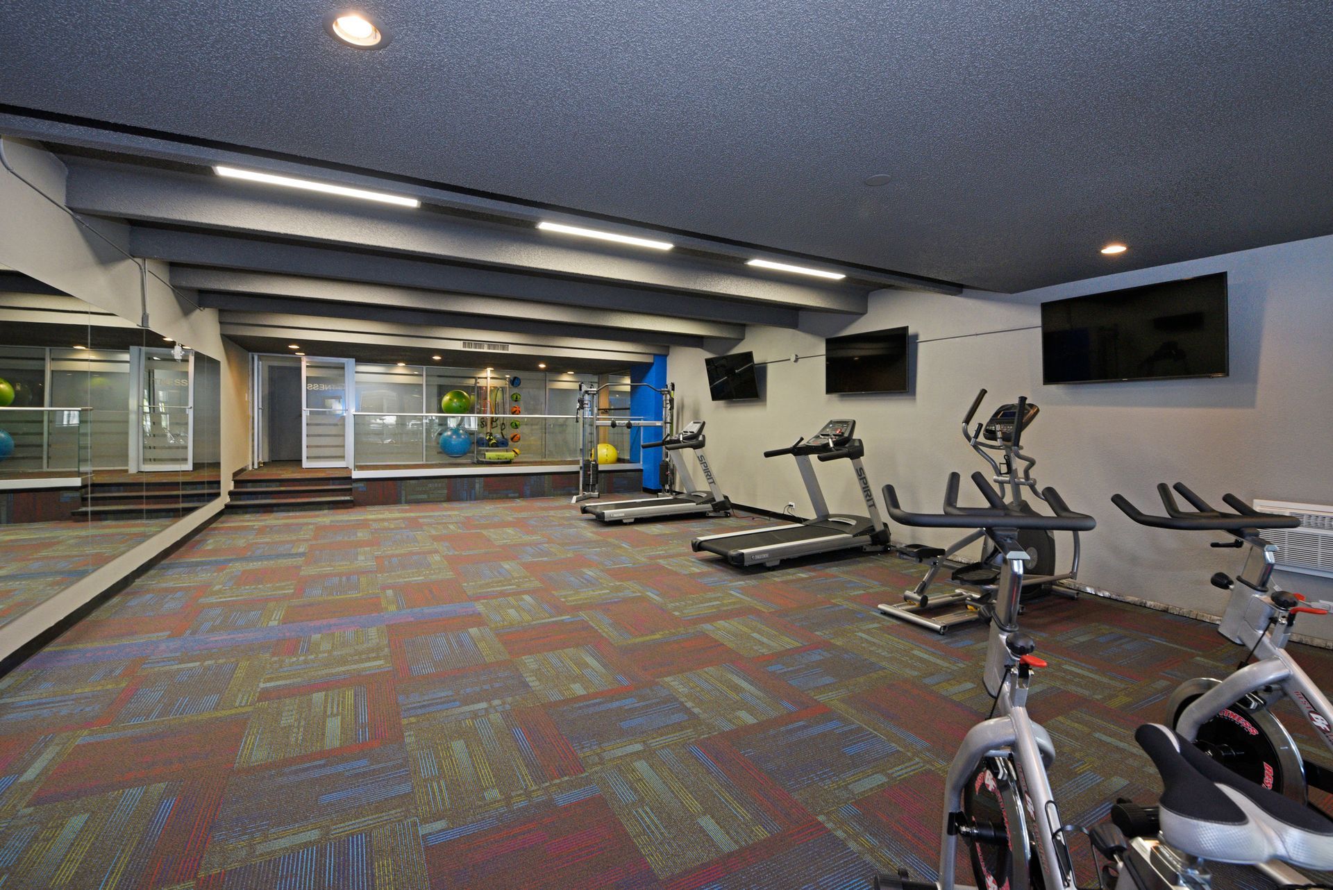 Empty gym with treadmills, exercise bikes, colorful carpet, and wall-mounted TVs.
