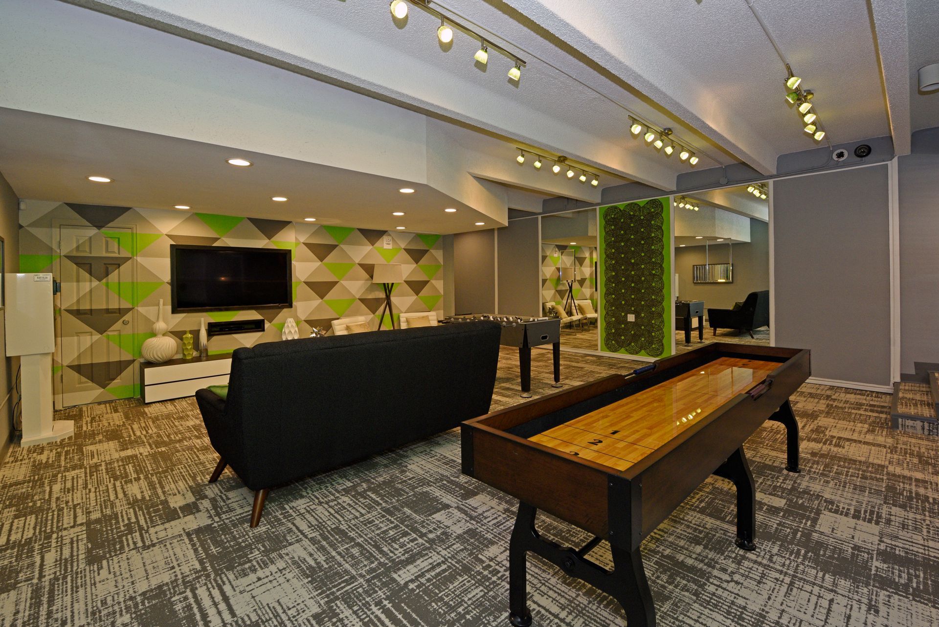 Modern lounge with sofa, wall TV, patterned carpet, and wooden shuffleboard table under recessed lights