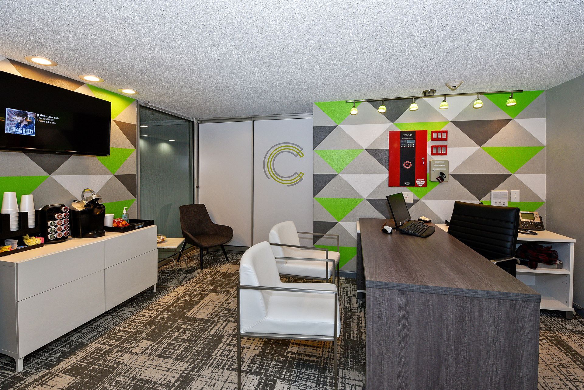 Modern office reception area with geometric wall art, white chair, dark desk, and TV screen.