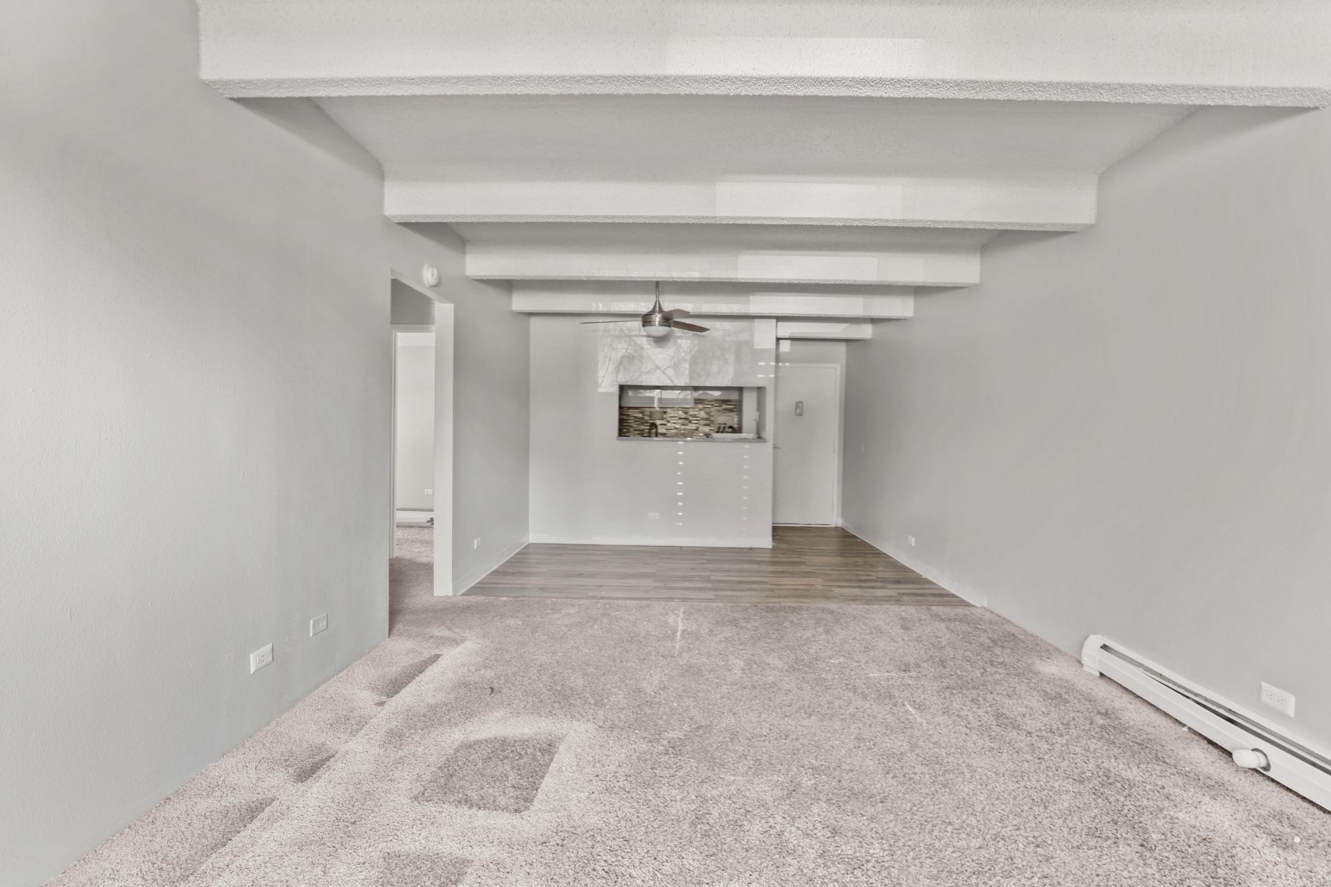 Empty carpeted basement room with white walls, exposed ceiling beams, and a small window at the far end