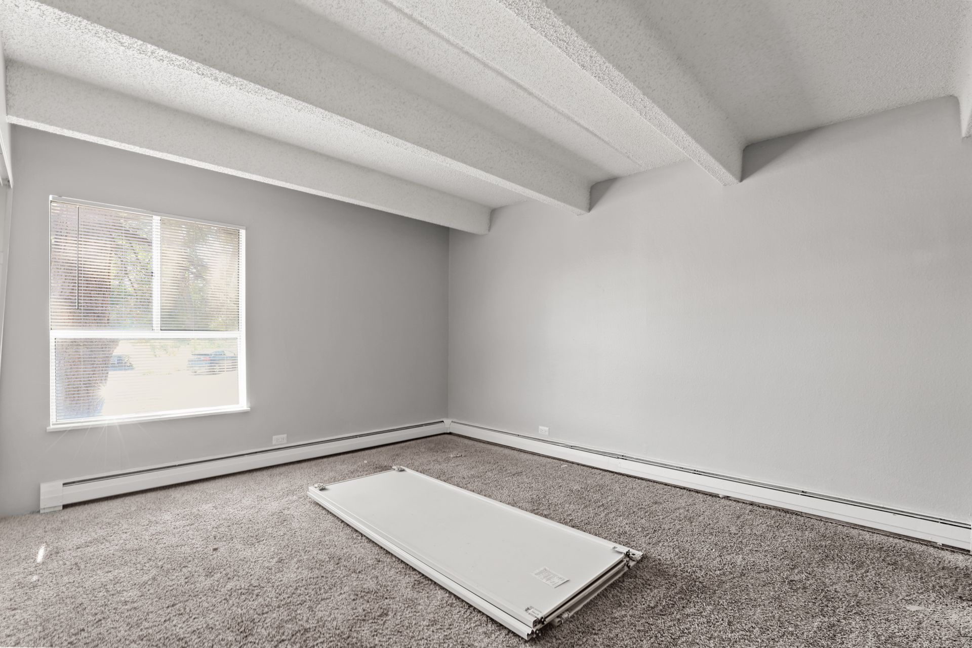 Empty gray bedroom with carpet, exposed ceiling beams, and a window with blinds.