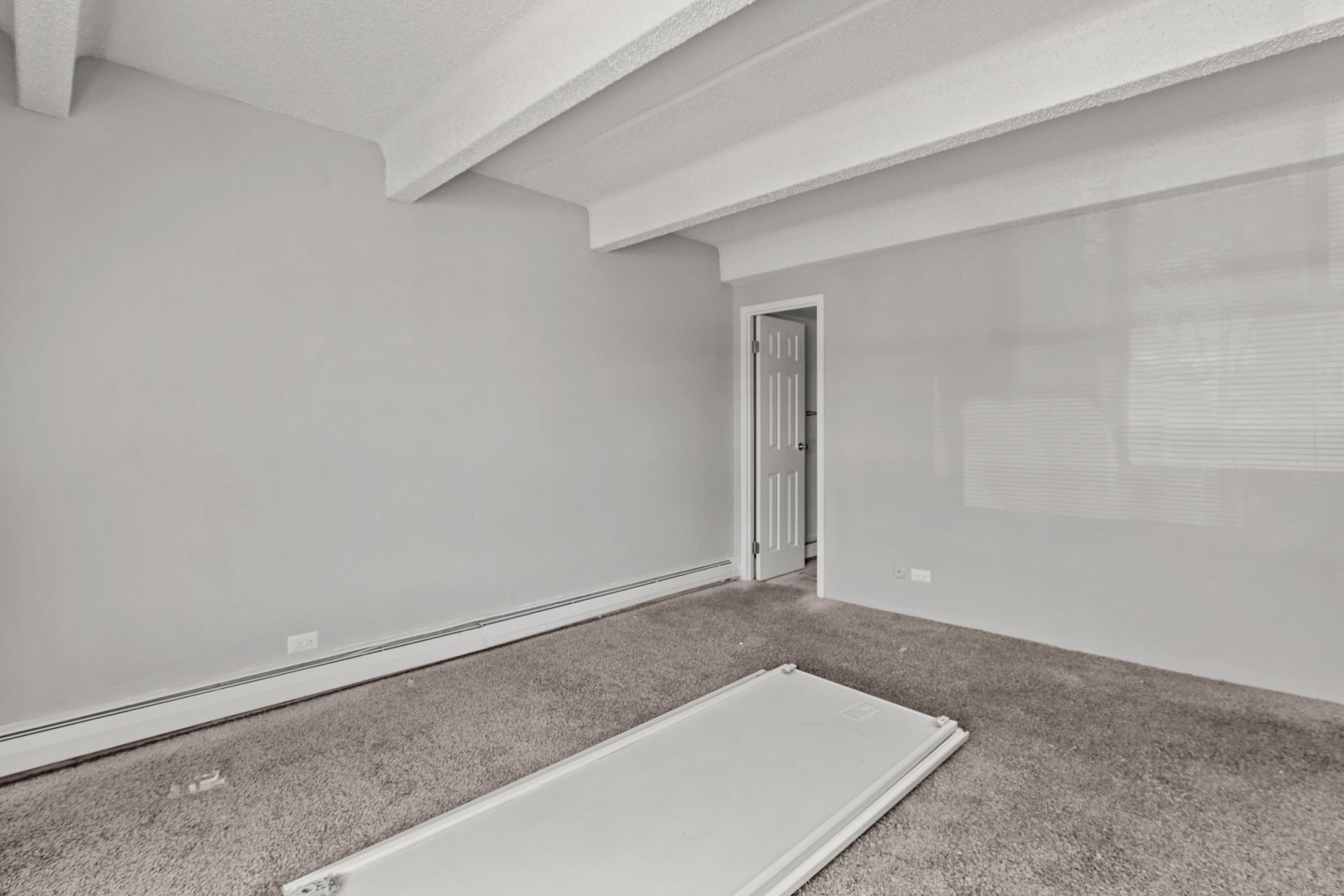Empty unfurnished room with gray carpet, white walls, and exposed ceiling beams.