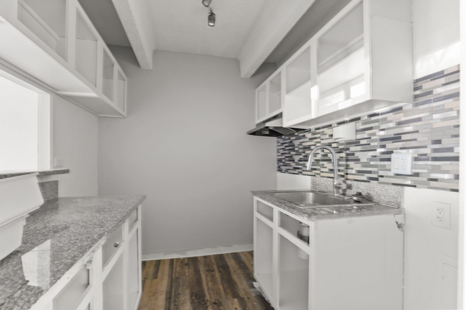 Bright white galley kitchen with gray countertops, mosaic backsplash, and dark wood flooring.