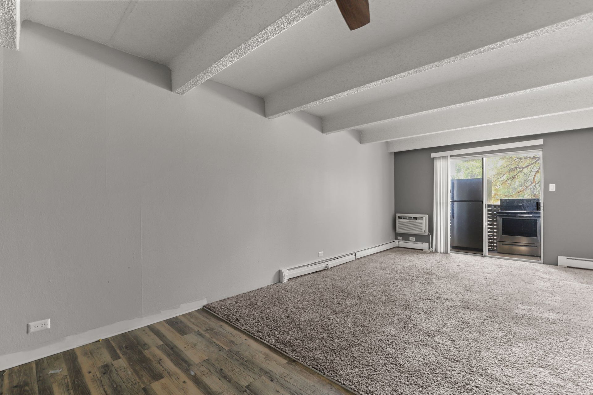 Empty carpeted living room with gray walls, exposed beams, and sliding doors to a balcony.