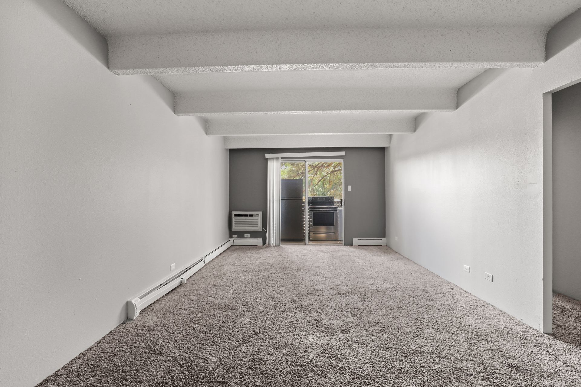 Empty carpeted living room with white walls, exposed ceiling beams, and a sliding glass door to a patio.
