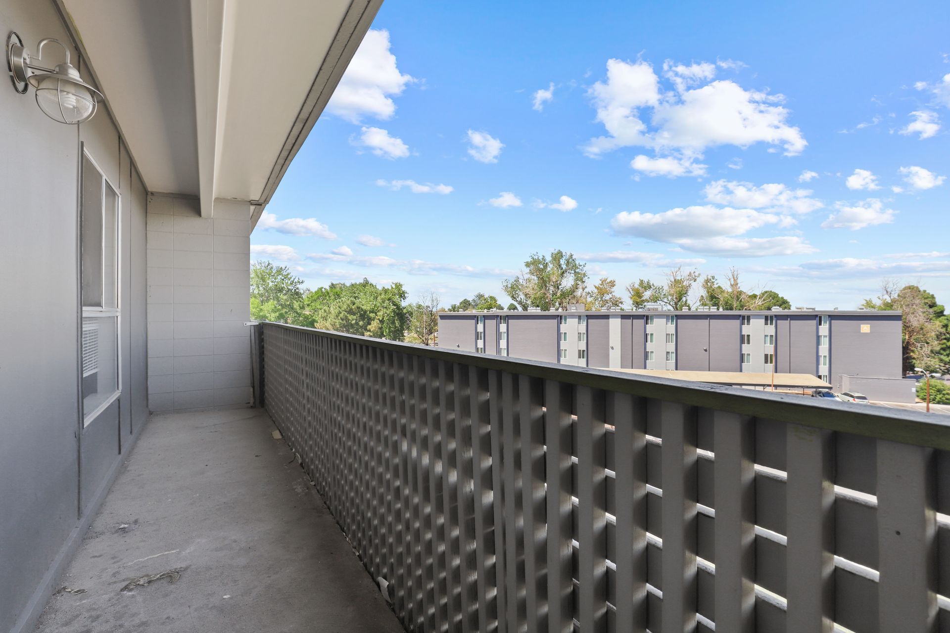 Apartment balcony with perforated railing, concrete floor, and blue sky over neighboring buildings