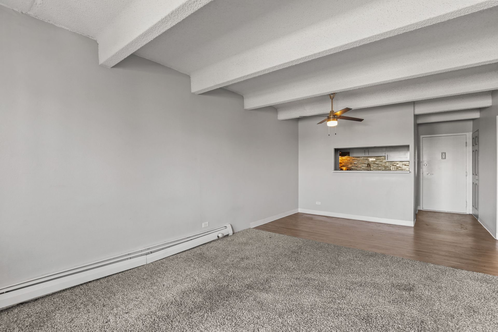 Empty carpeted living room with gray walls, white exposed ceiling beams, and wood flooring by a doorway