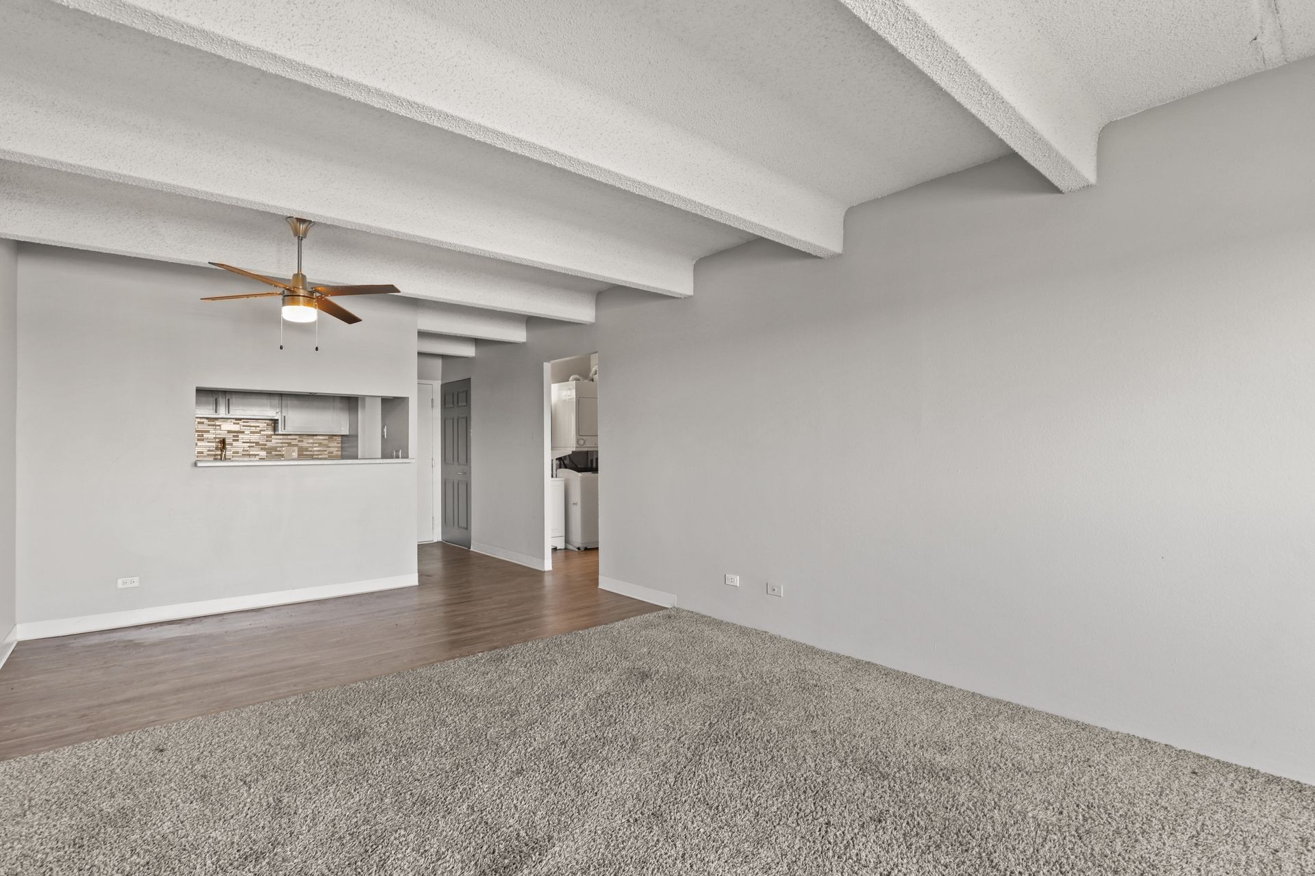 Spacious empty living room with gray carpet, wood floors, white beams, ceiling fan, and neutral walls