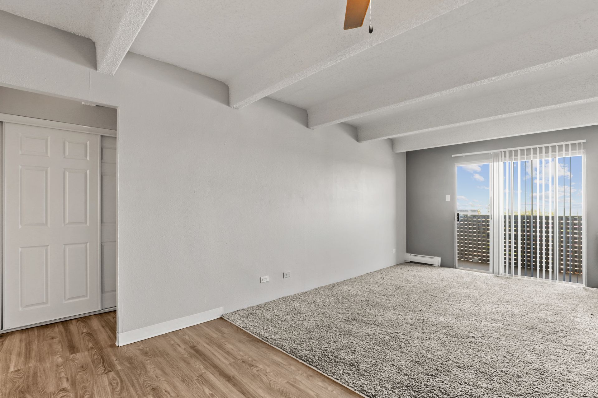 Empty modern bedroom with white walls, wood flooring, carpeted area, and sliding doors with vertical blinds.