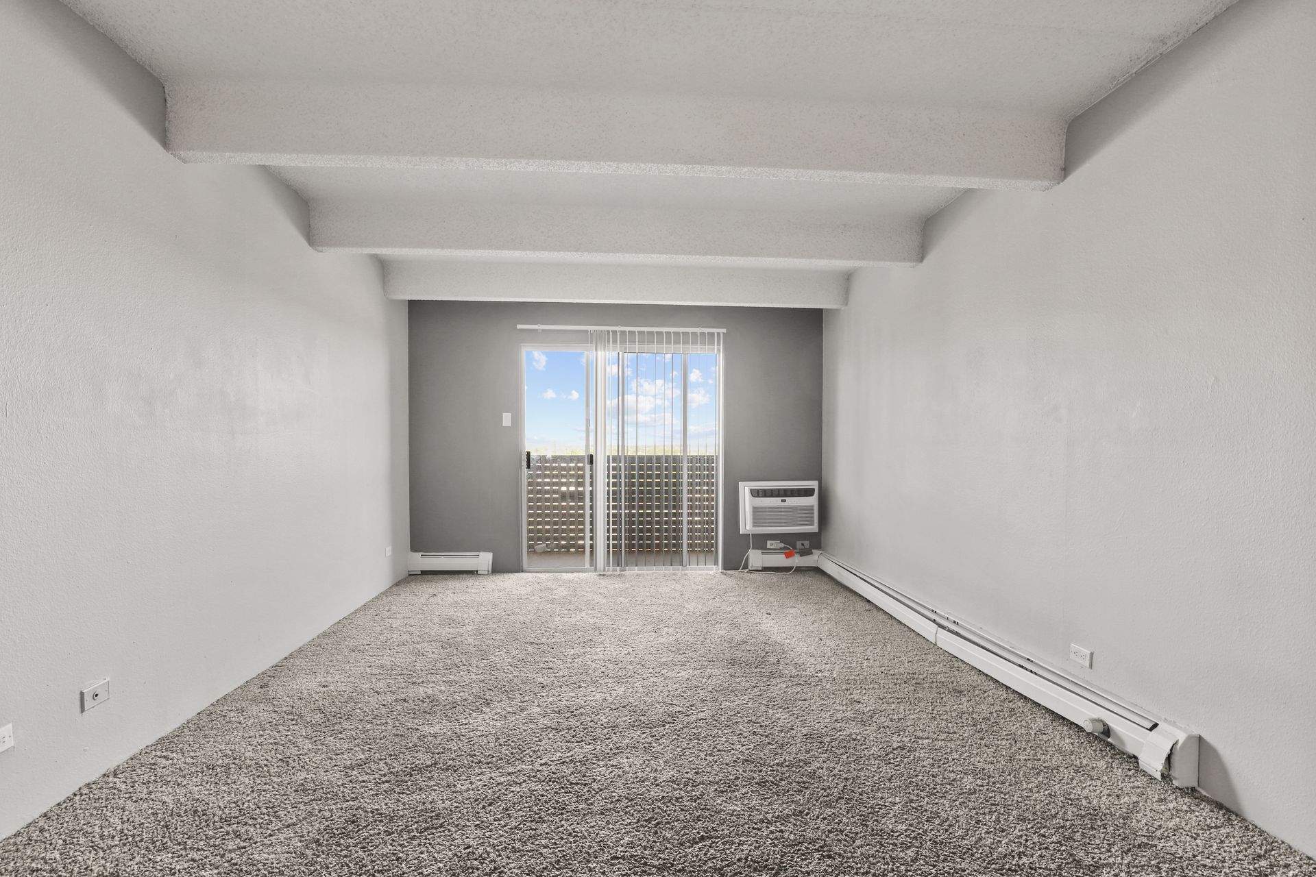 Empty carpeted living room with white walls, balcony doors, and a wall-mounted heater on the right.