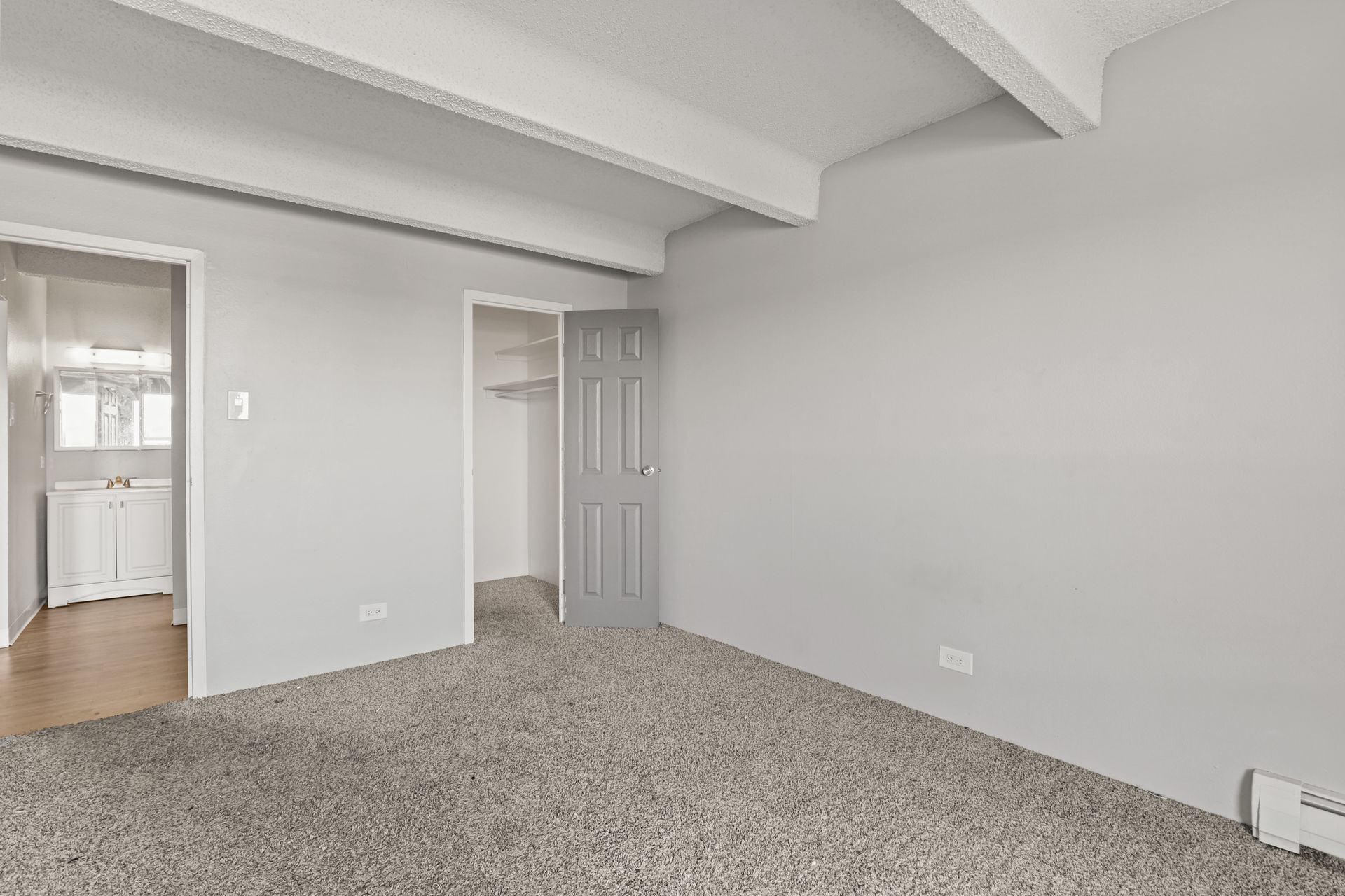 Empty carpeted room with white walls, exposed ceiling beams, and an open doorway to a kitchen.
