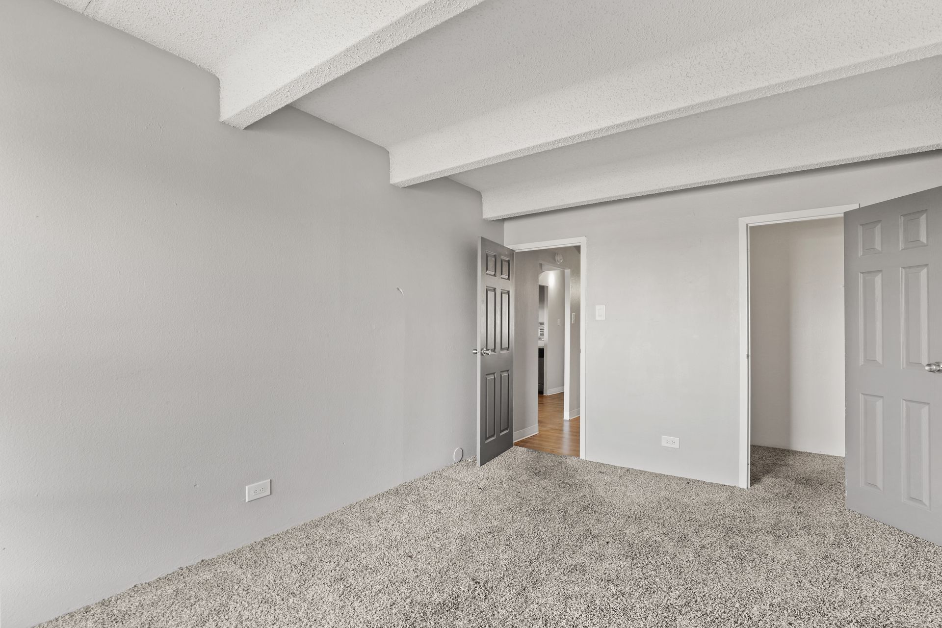 Empty carpeted bedroom with gray walls, white ceiling beams, and an open doorway.