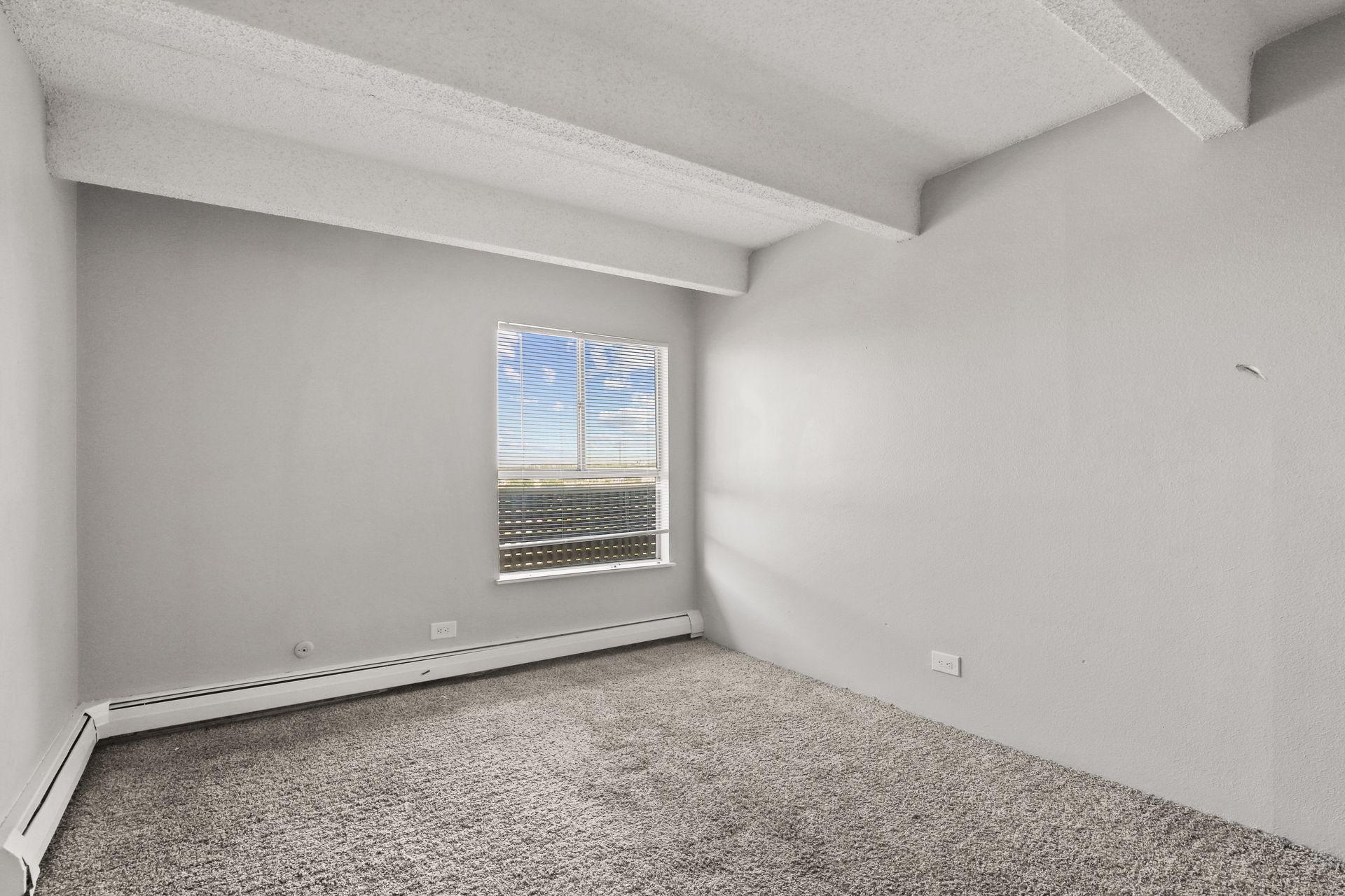 Empty white bedroom with carpet, exposed ceiling beams, and a small window with blinds.