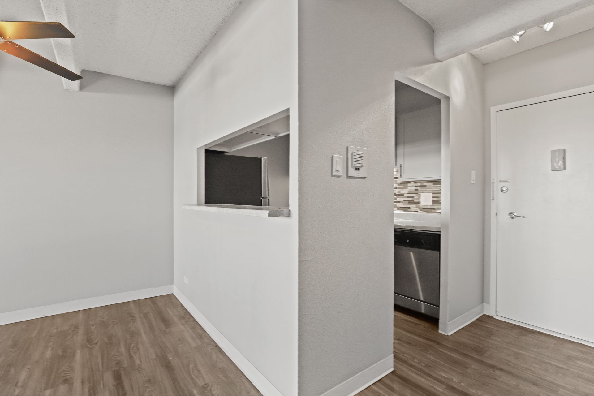 Empty white apartment room with wood floors, ceiling fan, and pass-through opening to the kitchen