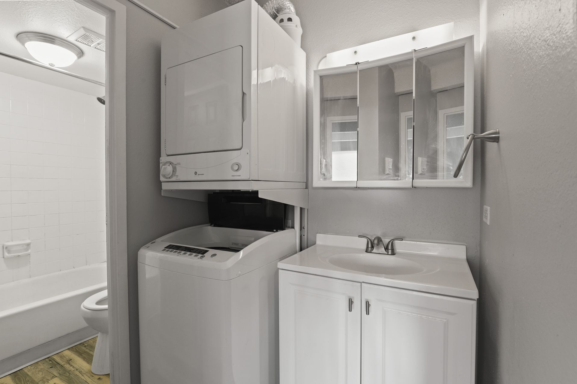 Small bathroom with bathtub, stacked washer and dryer, and white sink vanity with mirror cabinet.