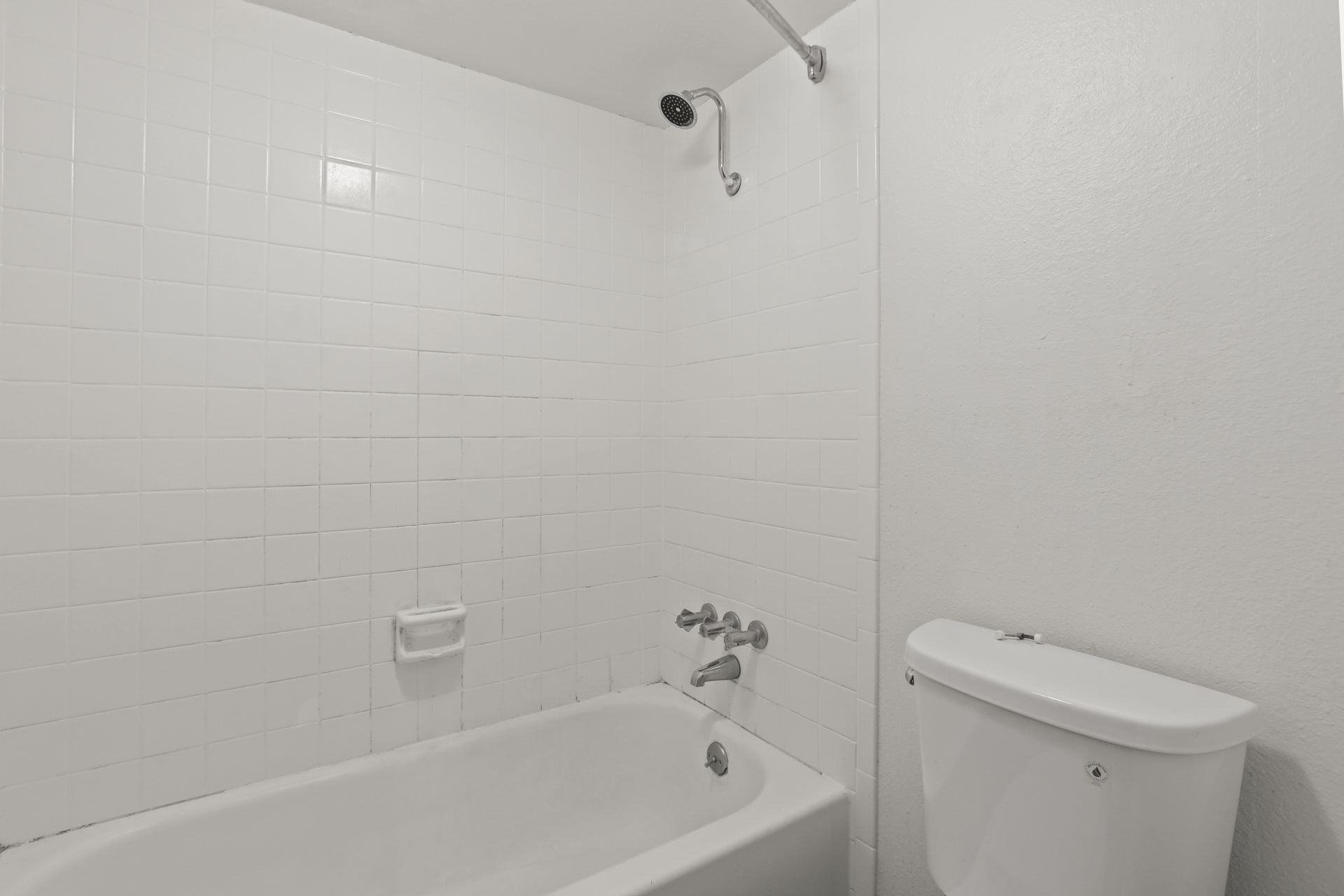Small white bathroom with a tub, toilet, tiled shower wall, and chrome fixtures.