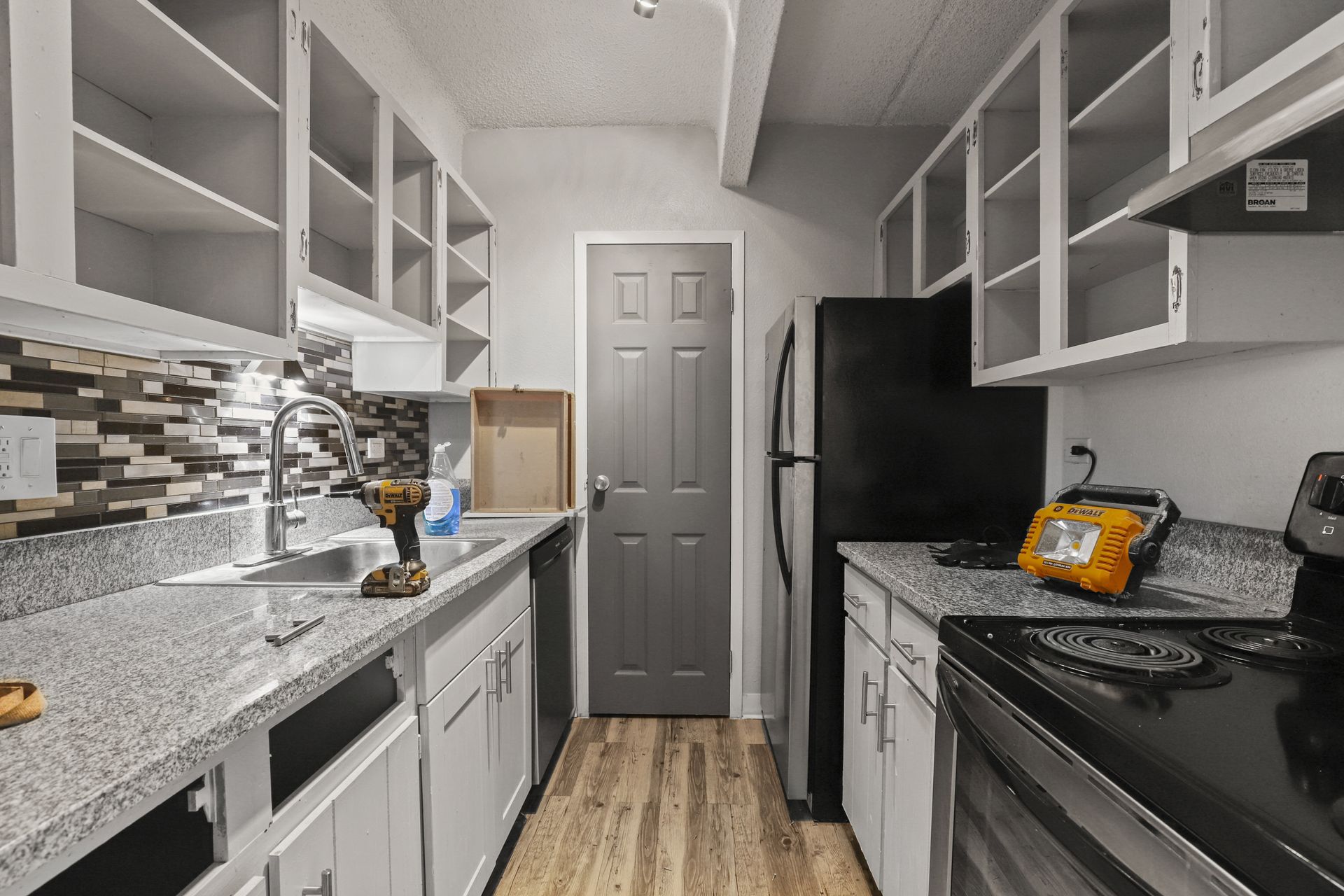 Narrow galley kitchen with white cabinets, speckled counters, black appliances, and a gray door at the end.