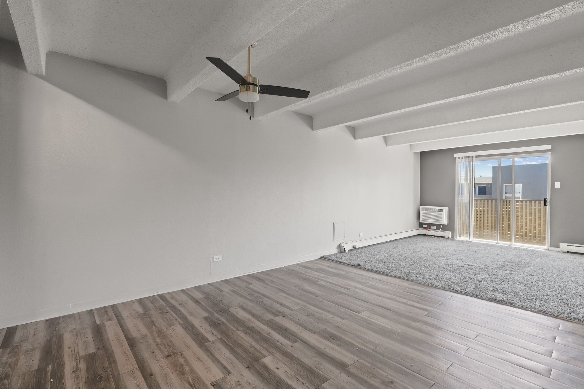 Empty modern living room with white walls, gray wood floors, exposed beams, and a ceiling fan.