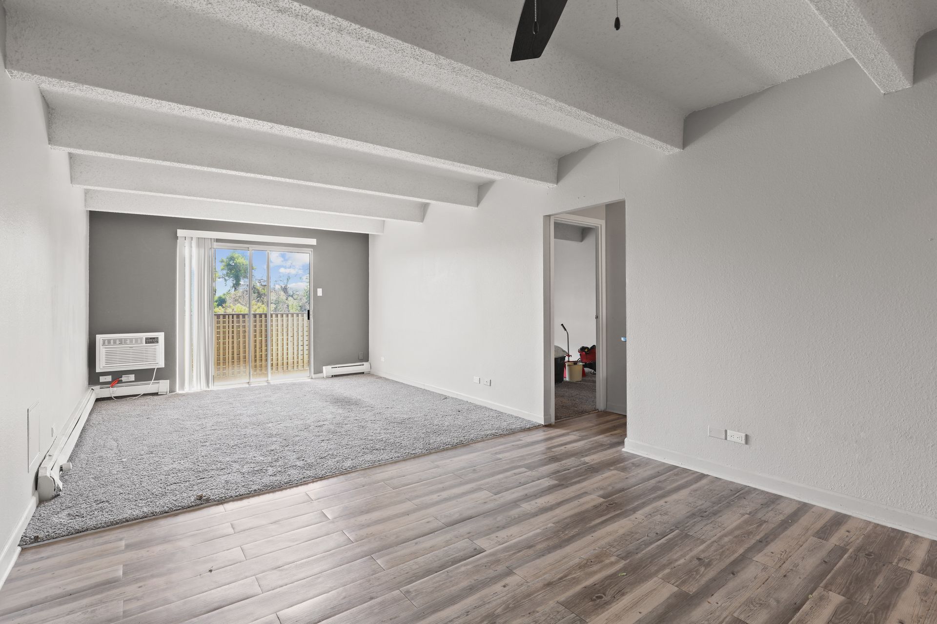 Empty living room with gray carpet, wood-look floor, white beams, and sliding door to a balcony.