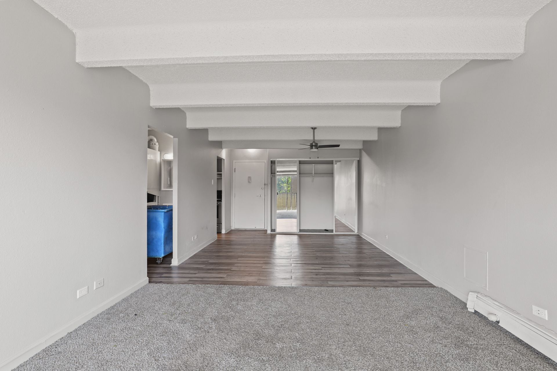 Empty apartment living area with gray carpet, wood floor, white walls, and exposed ceiling beams