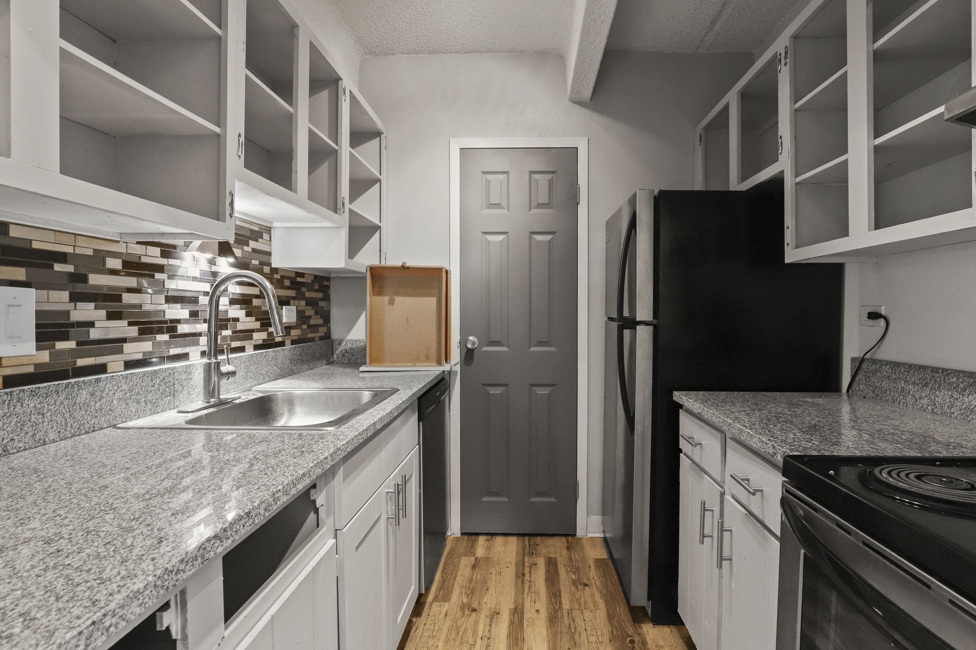 Narrow modern kitchen with white cabinets, speckled counters, black appliances, and a tiled backsplash.