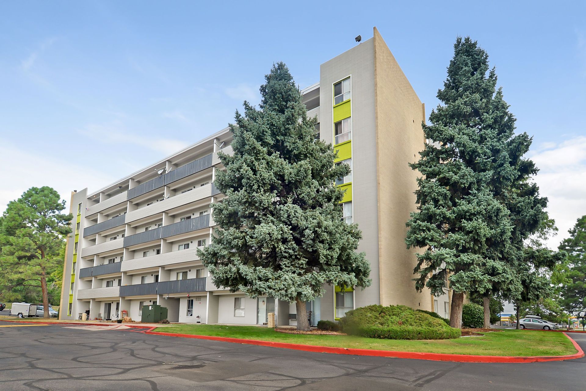 Apartment building exterior with balconies, tall evergreen trees, and a paved parking lot under a blue sky