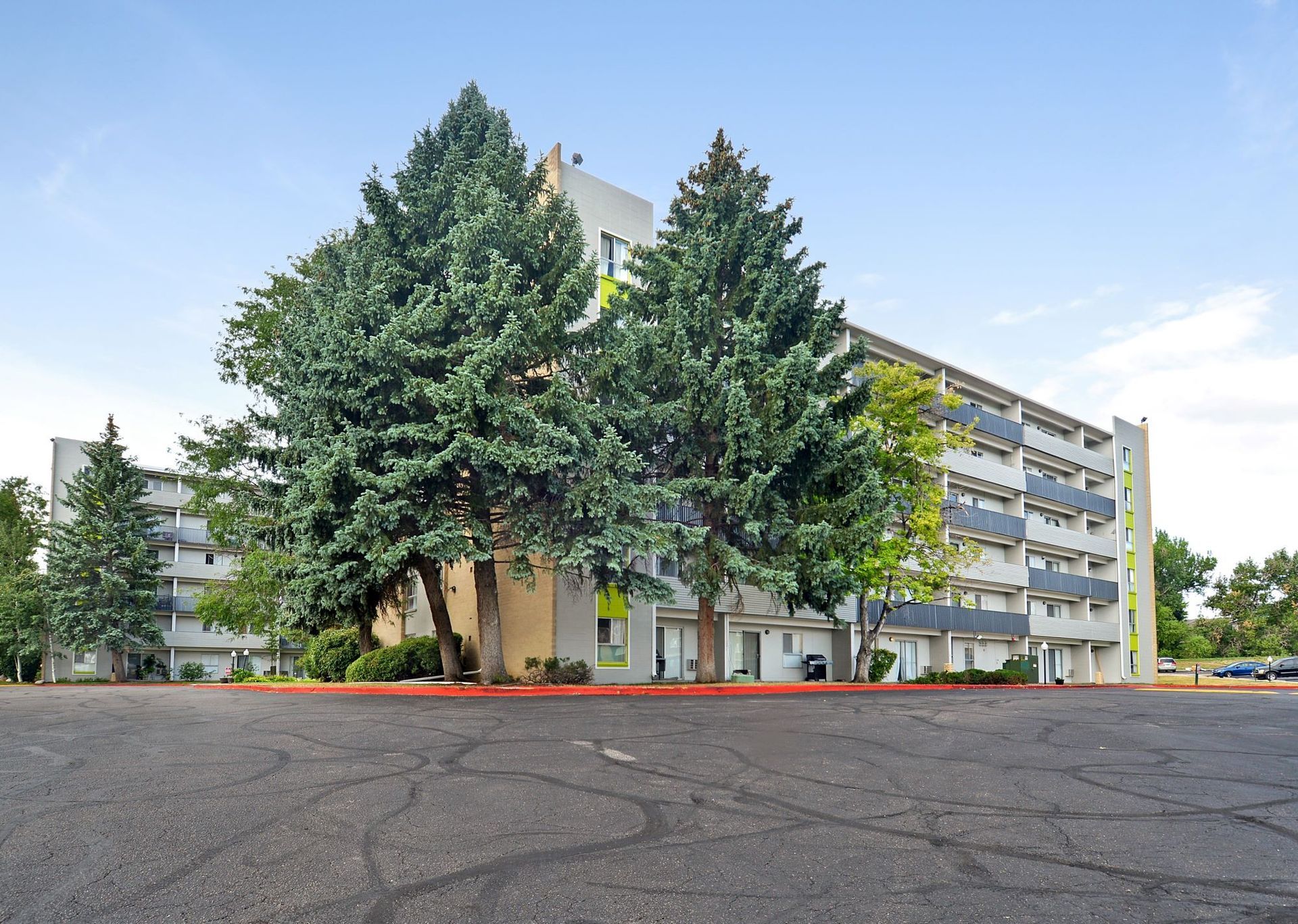 Apartment building behind large trees and a paved parking lot under a clear blue sky