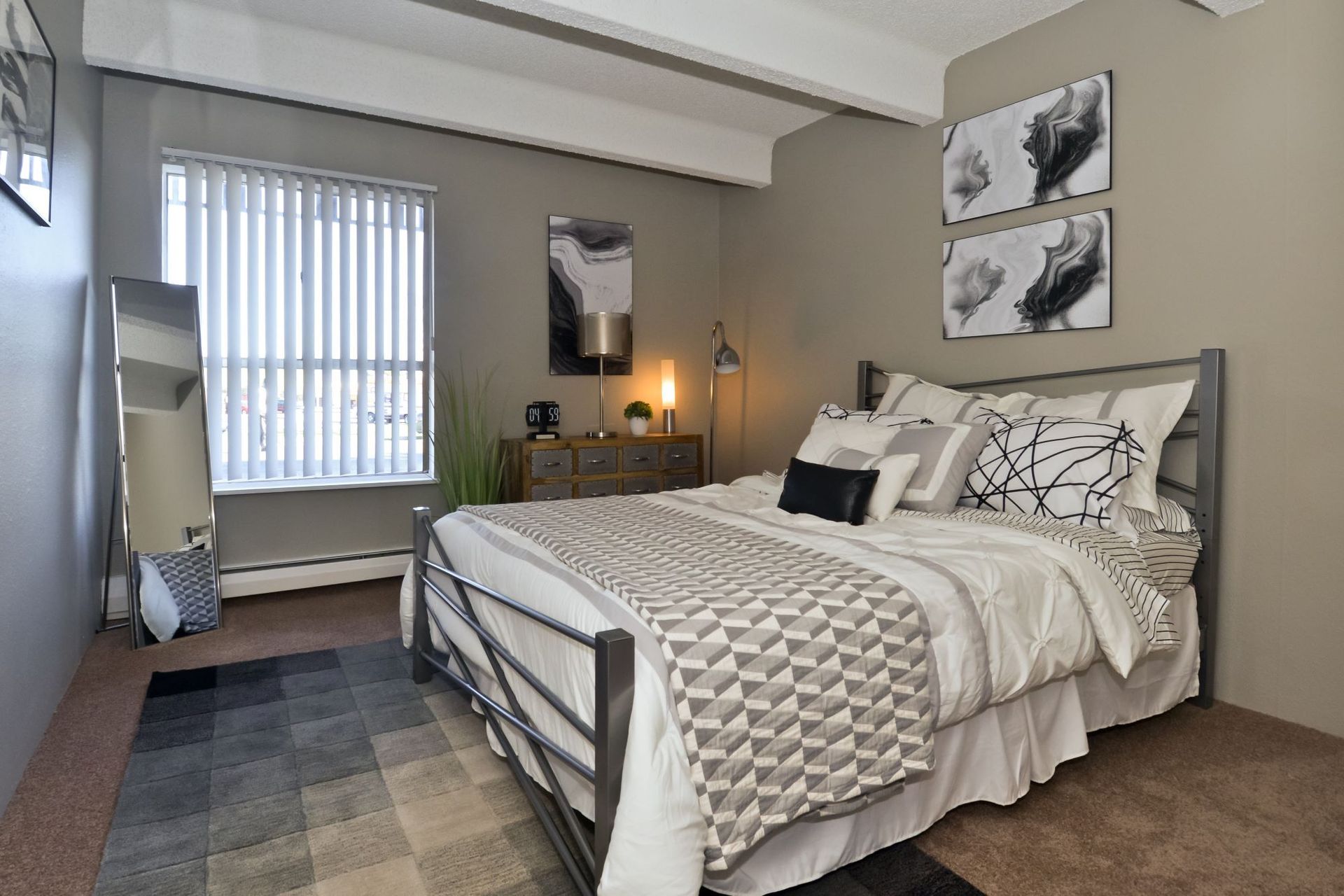 Neatly made bedroom with a gray bed, window, wall art, nightstand lamp, and full-length mirror