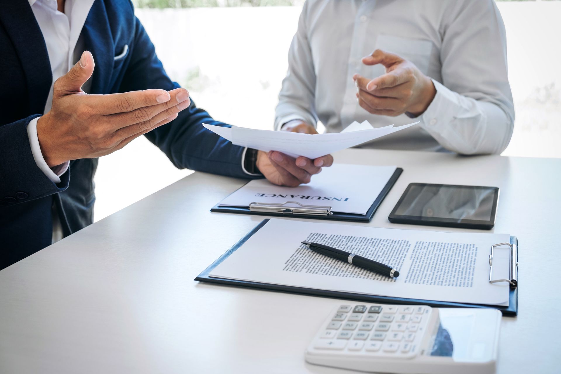 Business meeting discussing insurance documents with tablet and calculator on desk.