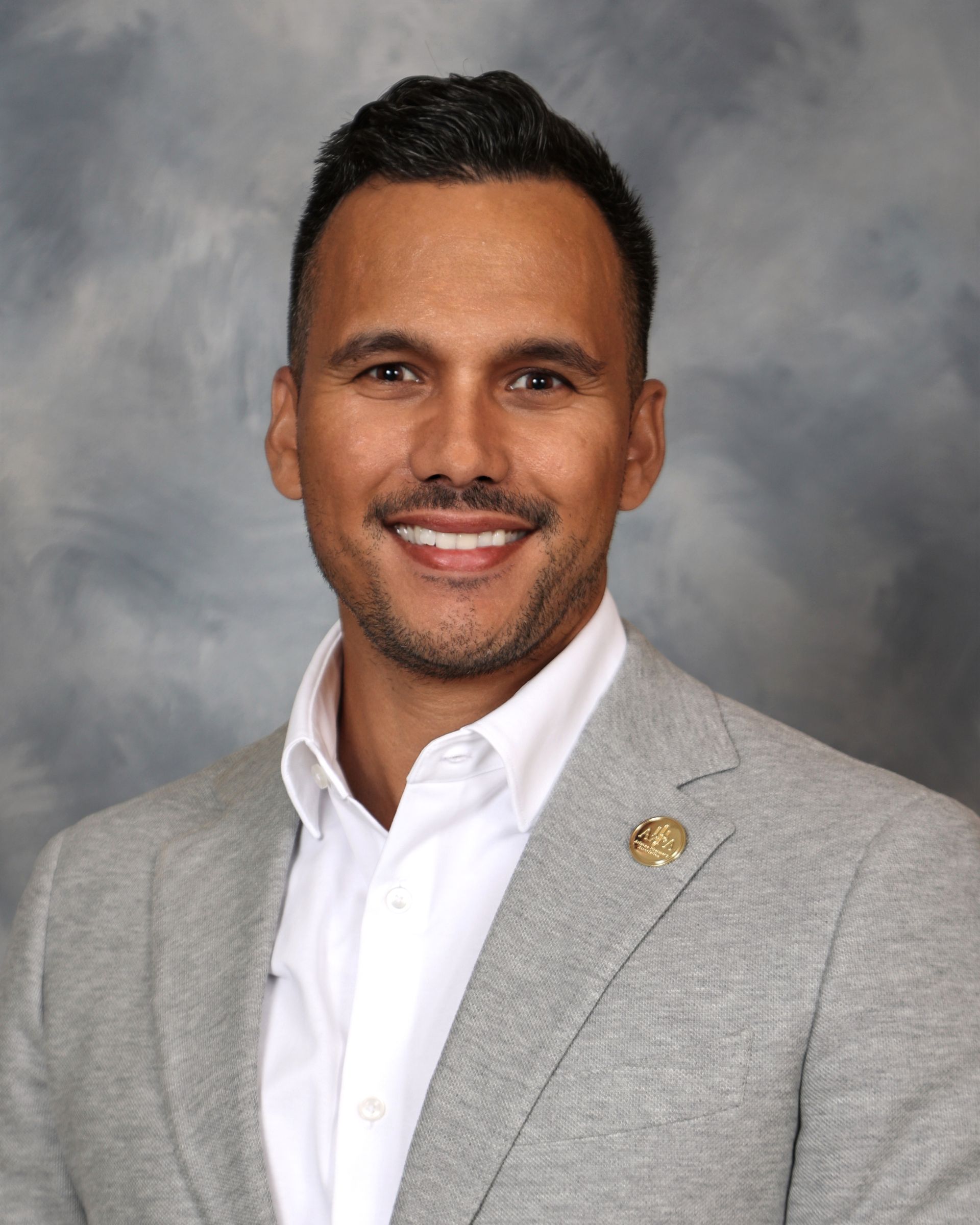 Man in grey blazer smiles, a white shirt beneath, against a blurred blue background.