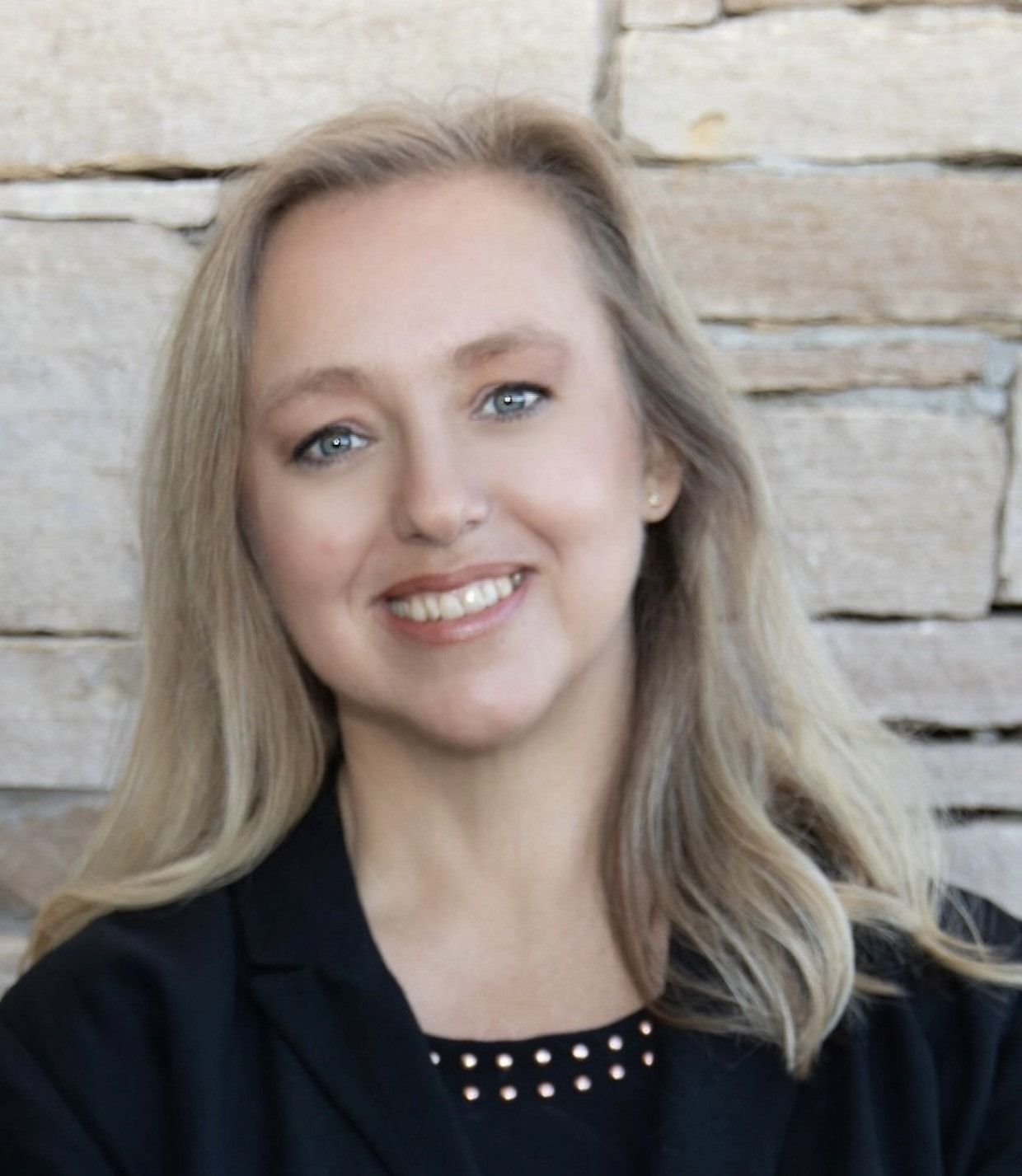 Blond woman smiling, wearing a black blazer, standing in front of a stone wall.