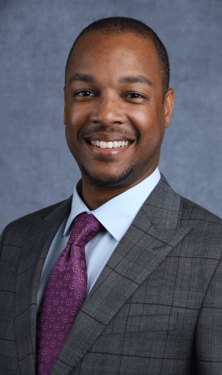 Man in a gray suit smiles, wearing a purple tie over a blue collared shirt.