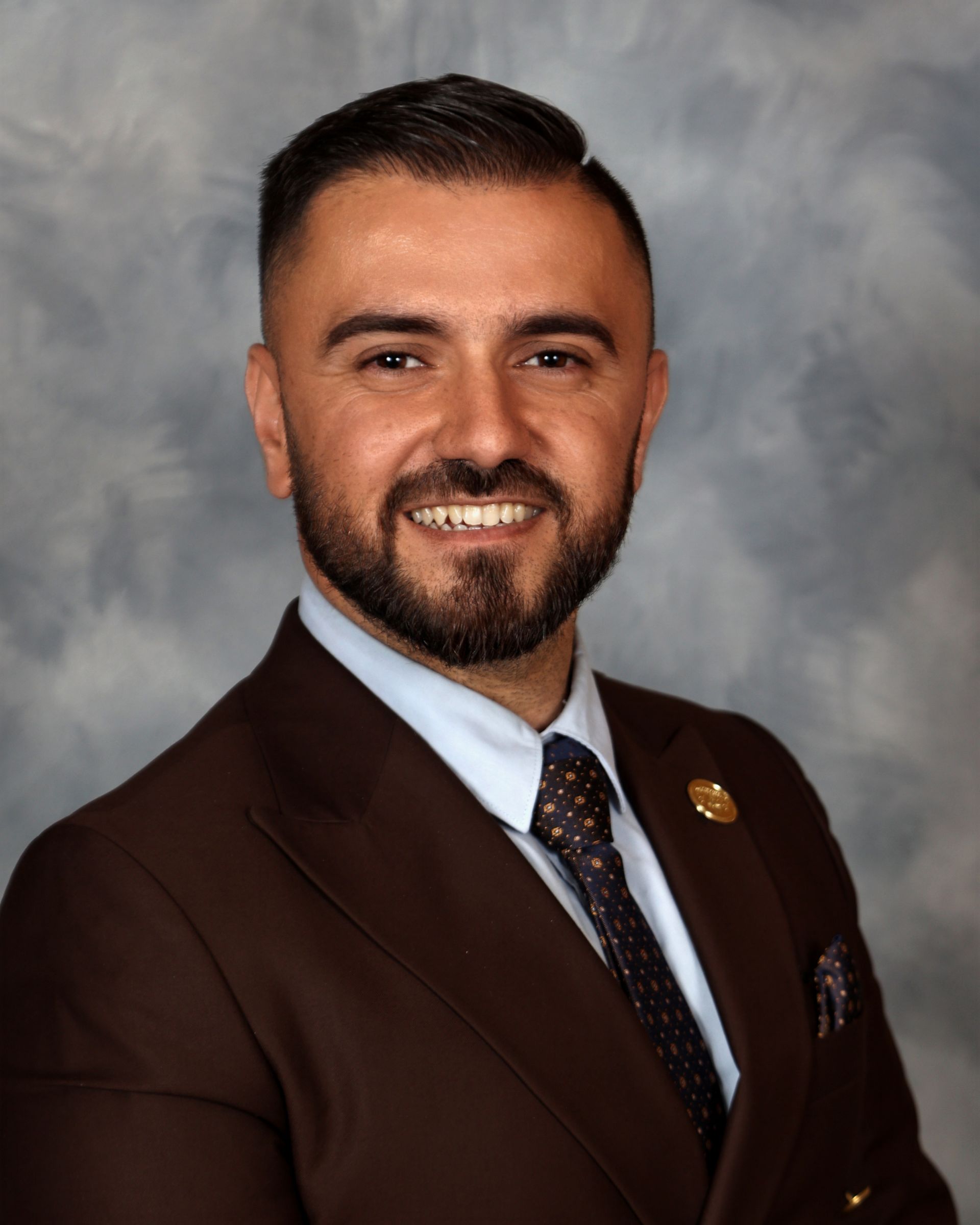 Man in suit jacket and purple shirt smiles; background is blue and gray.
