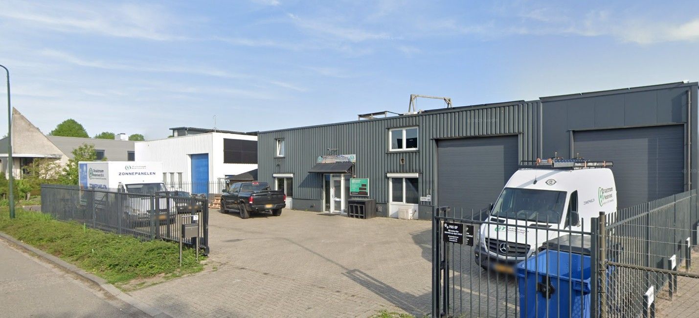 Industrieterrein met een magazijn en geparkeerde voertuigen achter een zwart hek op een zonnige dag.