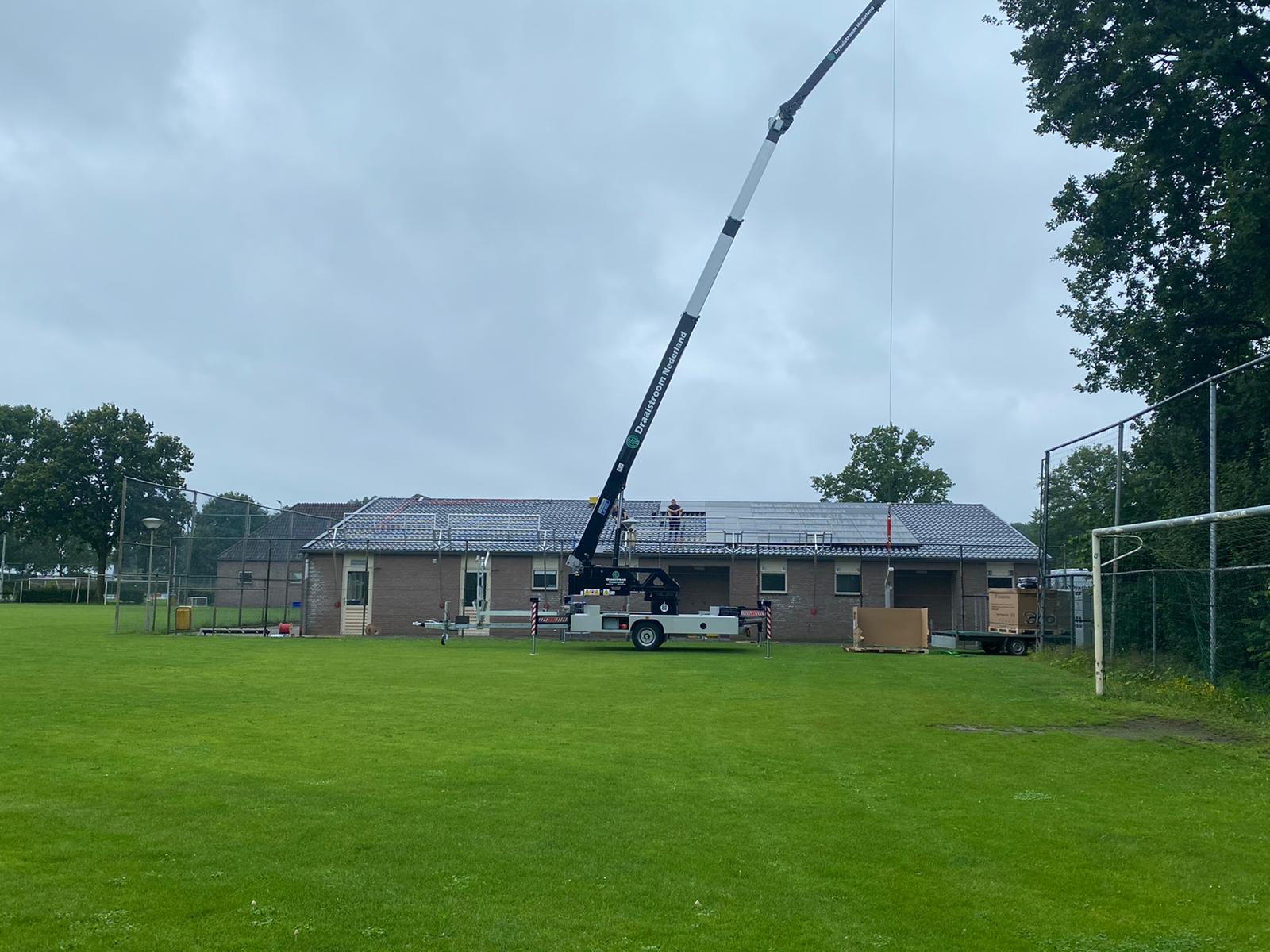 Een kraan hijst materialen naast een klein gebouw op een grasveld onder een bewolkte hemel.