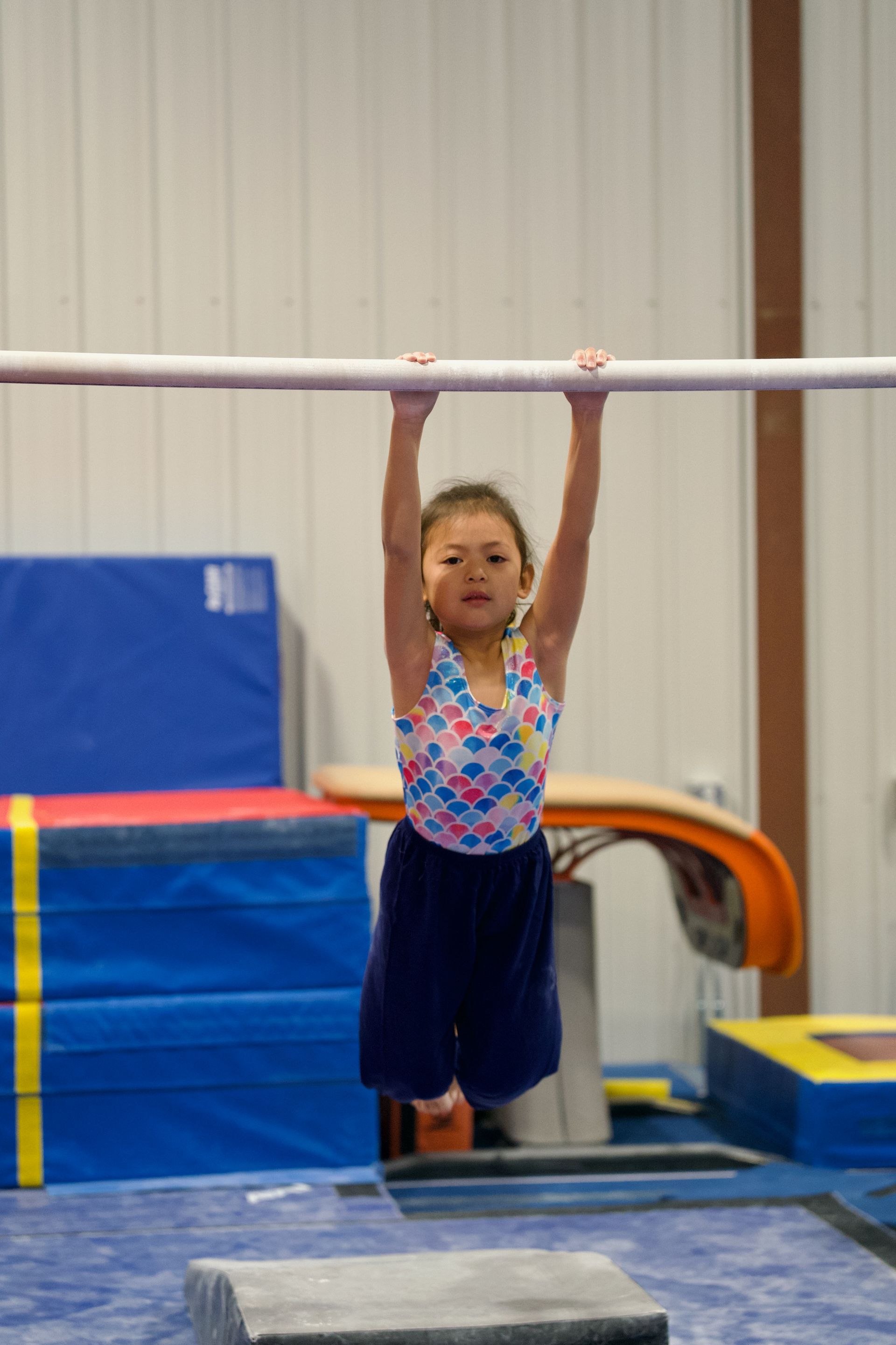 Child hanging from a horizontal bar in a gym, with colorful mats and equipment in the background.