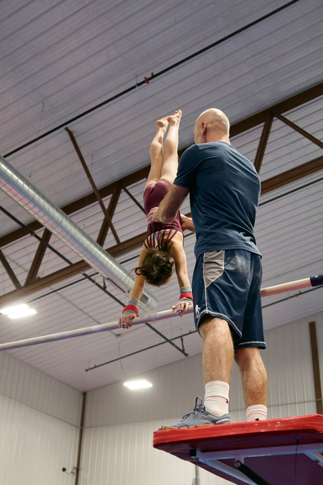 Gymnast hanging from the high bar in a gym, wearing red and blue leotard.