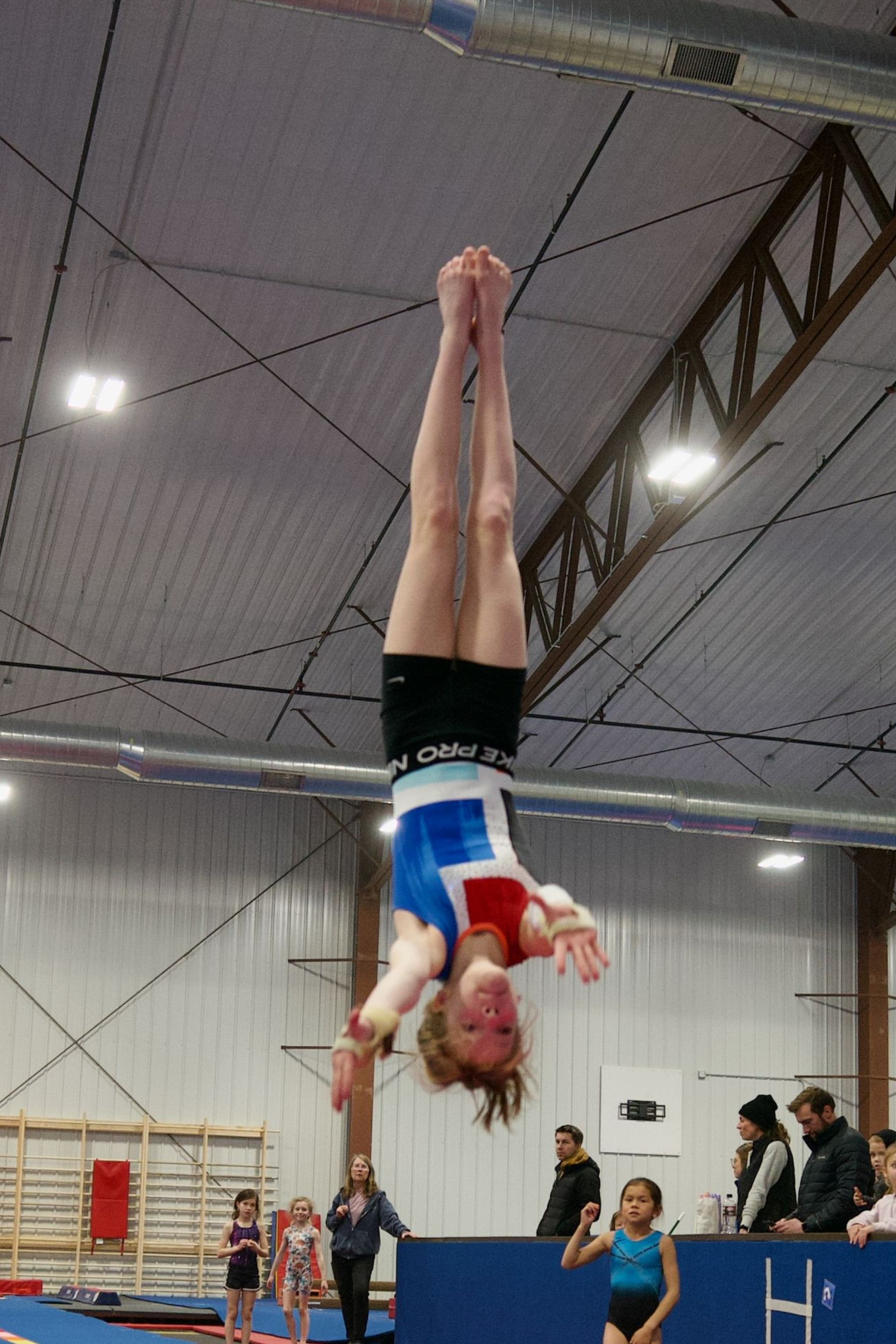 Gymnast in a black leotard balancing on a beam in a gym with colorful poles and mats