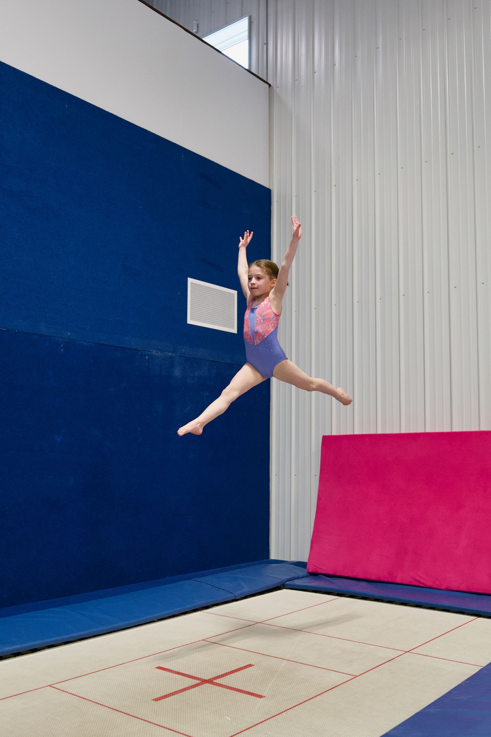Child midair during a gymnastics jump in a gym, above a marked trampoline or floor mat.