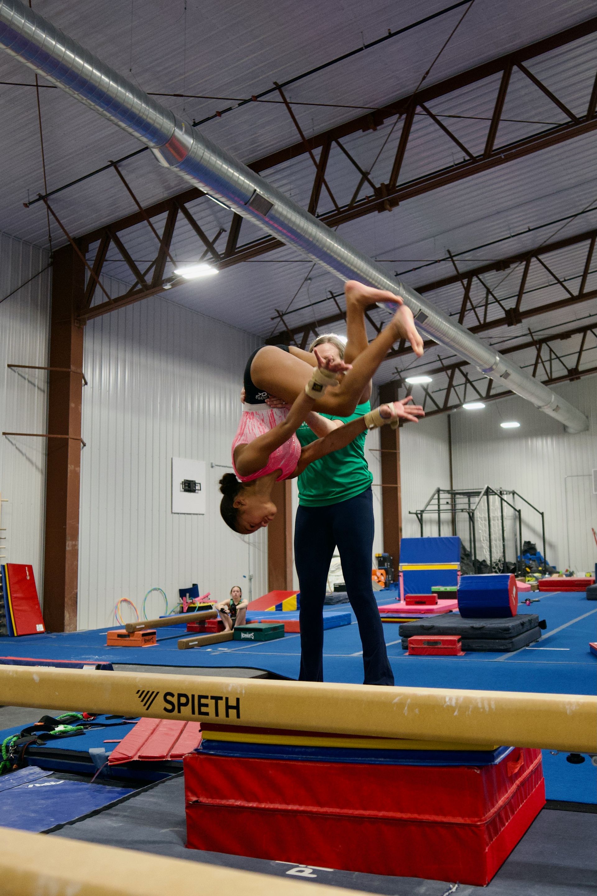 Gymnast doing a backbend on a balance beam with a coach spotting in an indoor gym.